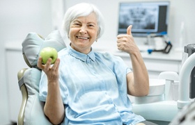 senior woman with all on four dental implants giving a thumbs up and holding an apple