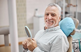 Older man with dental implants in DeLand smiling and holding mirror