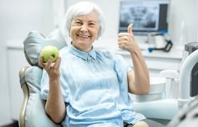 Woman holding apple gives thumbs-up after getting dental implant-retained dentures