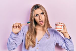 woman holding toothbrush in one hand and dentures in the other hand looking confused