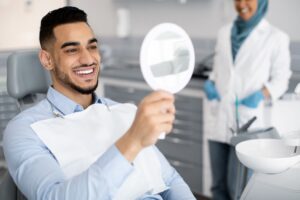 Man in blue shirt in dentist's chair smiling at his reflection in a handheld mirror