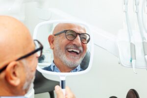 Older man with dental implants looking into handheld mirror in dentist’s chair and smiling.